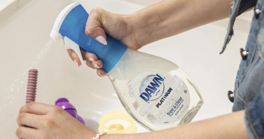 Hand spraying a bottle of Dawn Powerwash Free & Clear on dirty dishes in a sink