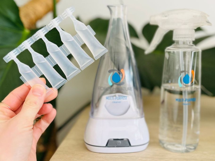 A Force of Nature Appliance and spray bottle with a human hand holding 5-capsules next t o them