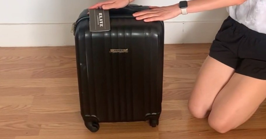 black hardside spinner suitcase with a woman sitting on her knees beside it, her hands on the top