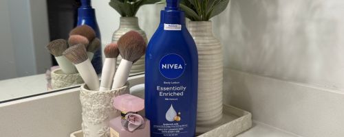 A bottle of Nivea Body Lotion in a bathroom tray with makeup brushes, perfume and a vase with flowers