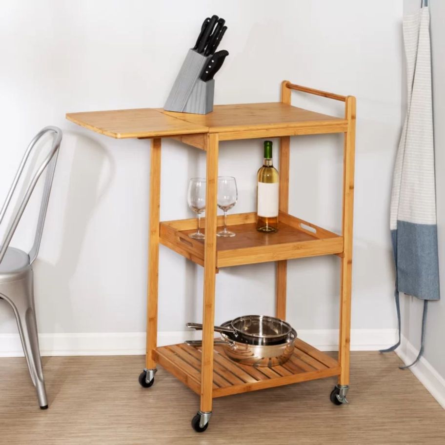 a natural wood colored rolling kitchen cart shown with kitchenware