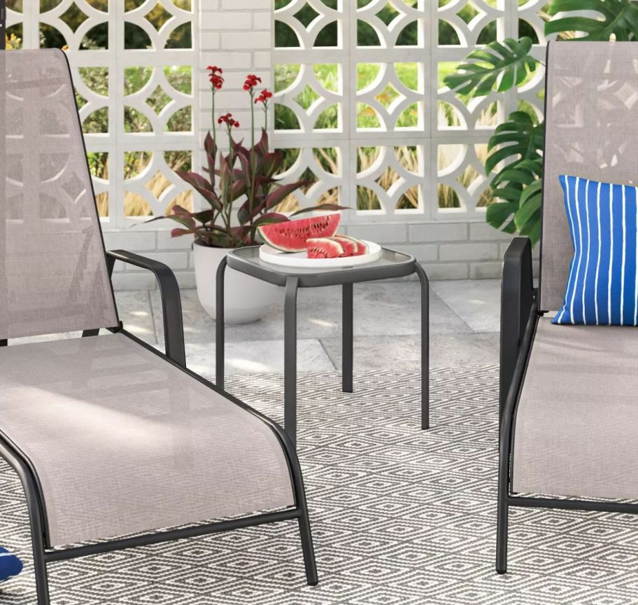 a glass top patio end table shown between two sling lounge chairs