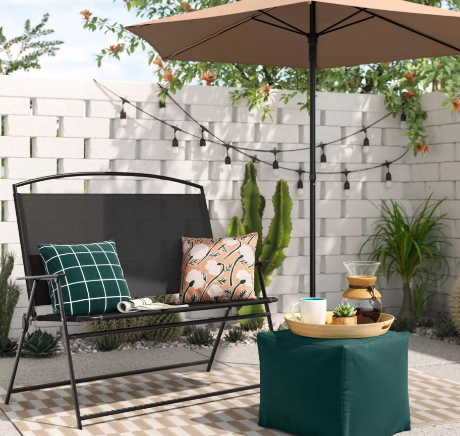 a 2 person beach chair on a patio shown with an umbrella and a green pouf