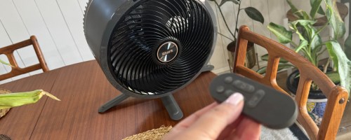 hand holding a remote control pointed to Shark fan that is sitting on top of the table
