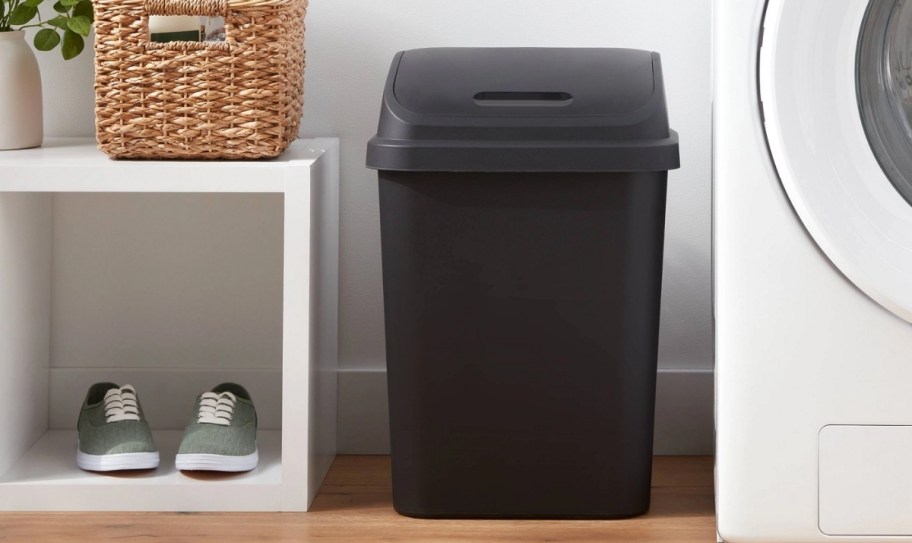 a black plastic flip top trash can next to a washing machine