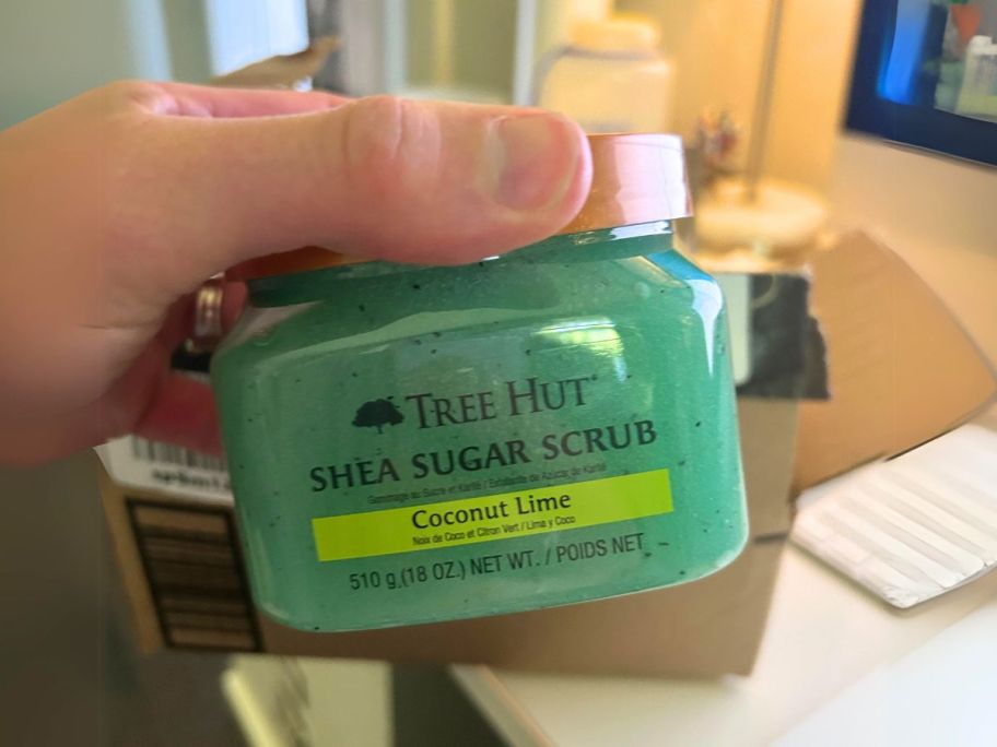 hand holding Tree Hut Shea Sugar Body Scrub in Coconut Lime 18oz in front of Amazon box