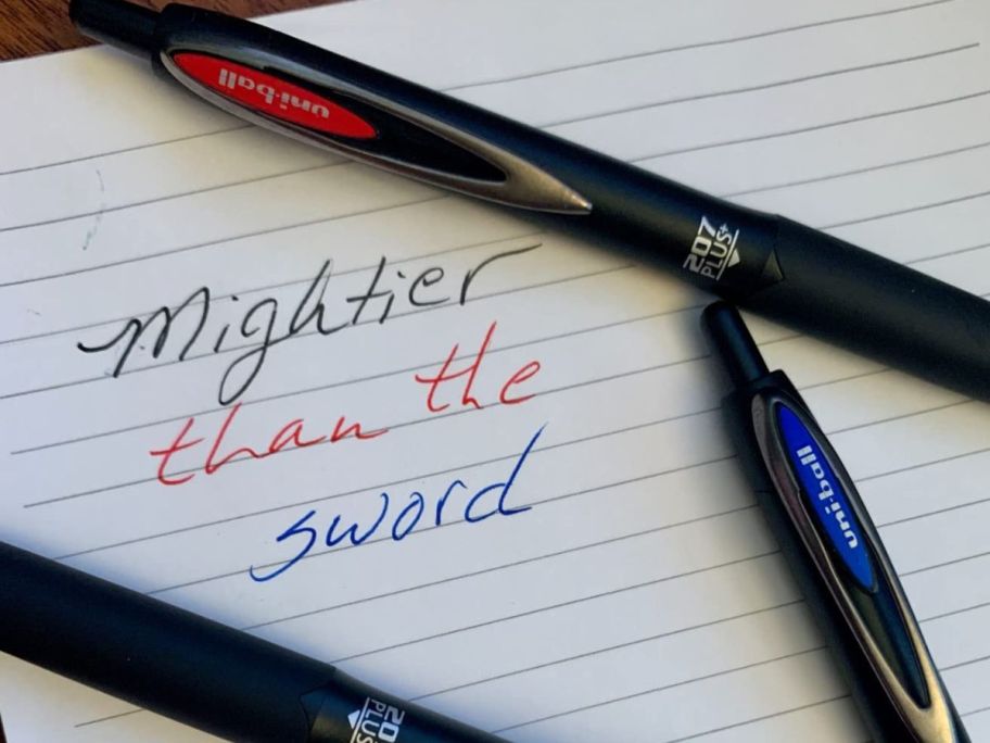 A paper with 3 Uniball colored pens with Mightier Than the Sword written in black blue and red ink