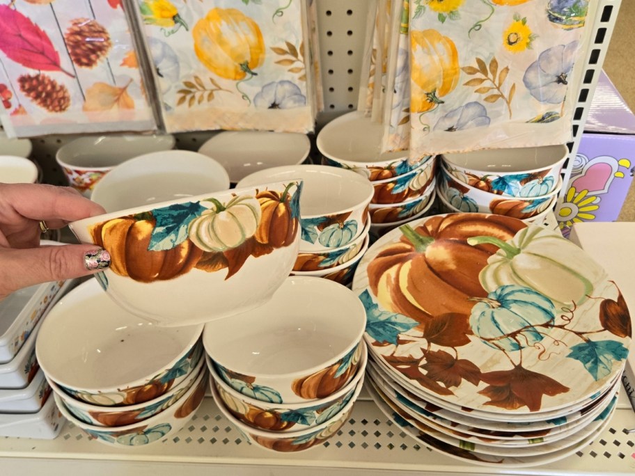 hand holding a dinnerware bowl with pumpkins on it, more bowls and dishes behind it