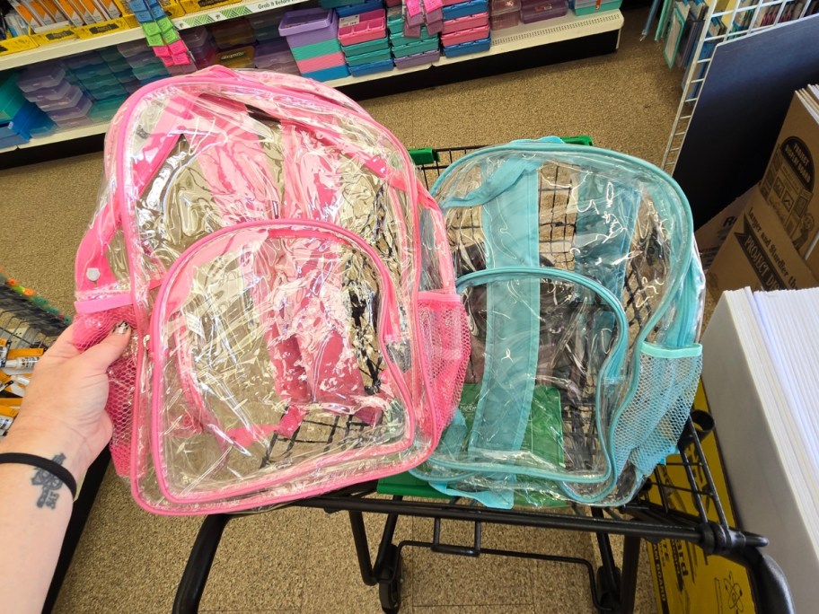 hand holding a pink and clear kid's backpack next to a blue and clear one in a cart