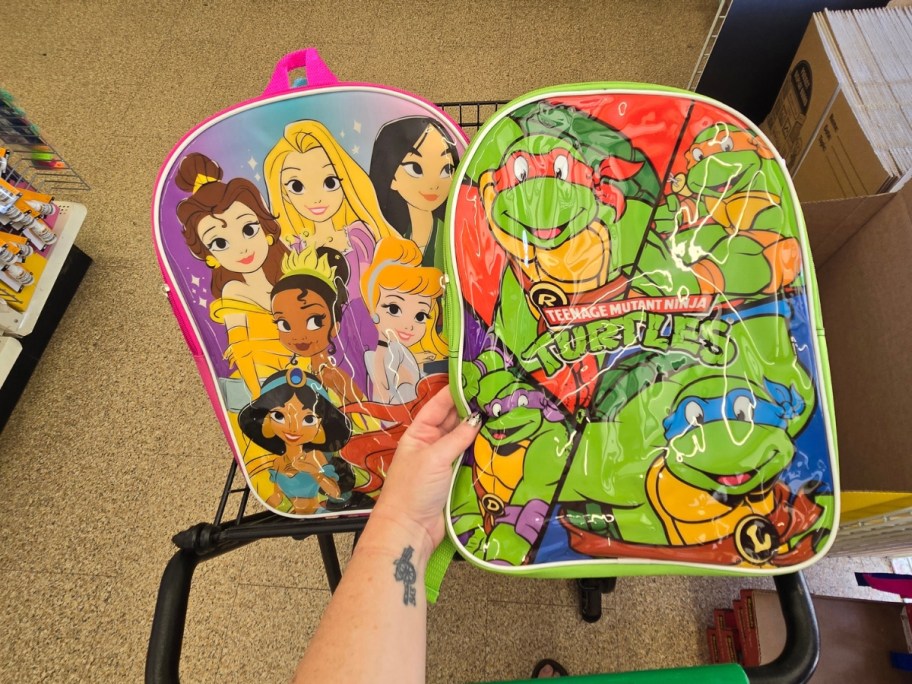 hand holding a Teenage Mutant Ninja Turtles kid's backpack next to a Disney Princess kid's backpack in a cart