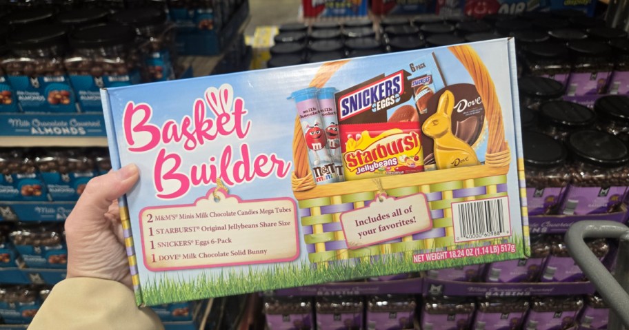 A hand holds a colorful "Basket Builder" box in a store aisle. The box features chocolates and candies like M&M's, Snickers, Starburst, and a Dove chocolate bunny, set in an illustrated basket. The background shows shelves filled with other products.