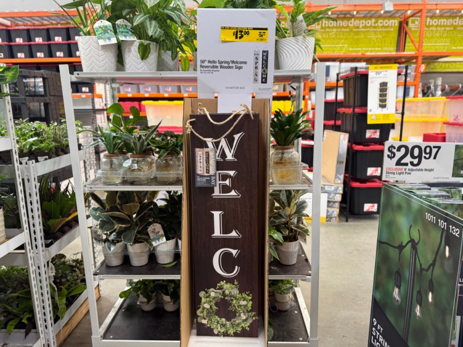 A tall, vertical wooden "Welcome" sign with a yellow $13.00 clearance tag on a store display shelf surrounded by potted plants.
