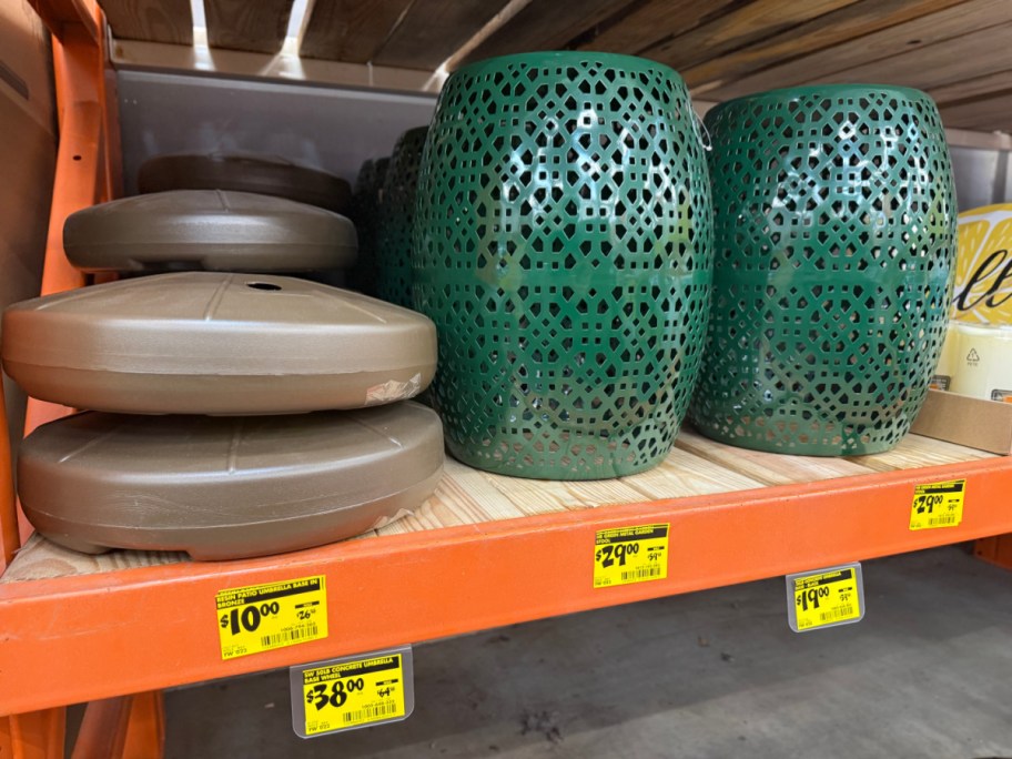 A stack of brown patio umbrella bases and two green cutout metal garden stools on an orange store shelf above yellow clearance tags.