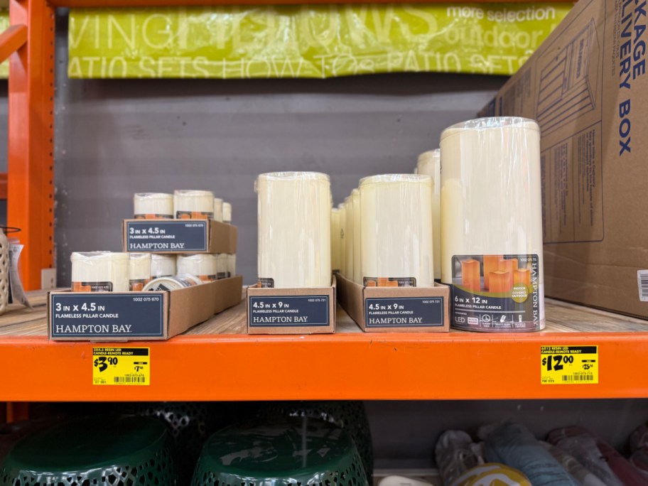 Various sizes of Hampton Bay flameless pillar candles in display boxes on an orange store shelf above yellow clearance tags.