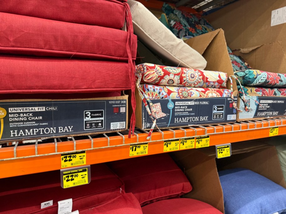 Stacks of solid red and floral Hampton Bay outdoor chair cushions on an orange store shelf above yellow clearance tags.