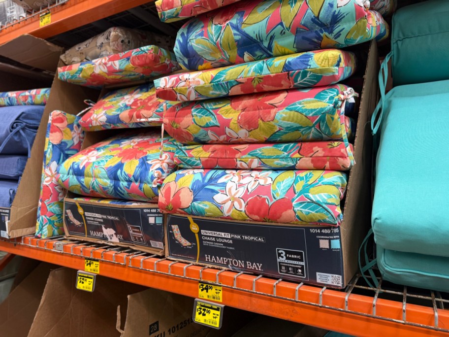 Stacks of pink tropical floral and solid teal Hampton Bay outdoor chaise lounge cushions on an orange store shelf above yellow clearance price tags.