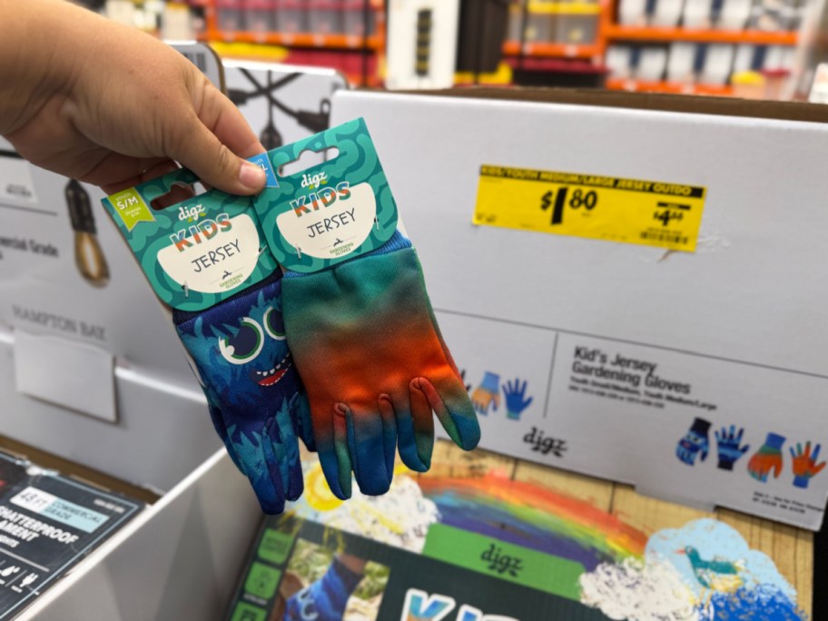 A hand holding two pairs of Digz kids' gardening gloves, one with a blue monster design and the other tie-dye, in front of a store display box featuring a yellow $1.80 clearance tag.