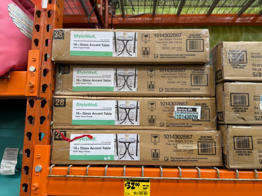 A stack of four cardboard boxes for StyleWell 18-inch glass accent tables on an orange store shelf above a yellow $32.00 clearance tag.