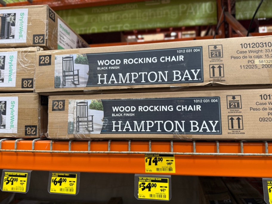 Two cardboard boxes for Hampton Bay black wood rocking chairs on an orange store shelf above yellow clearance price tags.