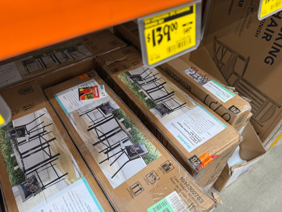 Cardboard boxes for StyleWell 3-piece patio dining sets stored under an orange store shelf beneath a yellow $139.00 clearance tag.