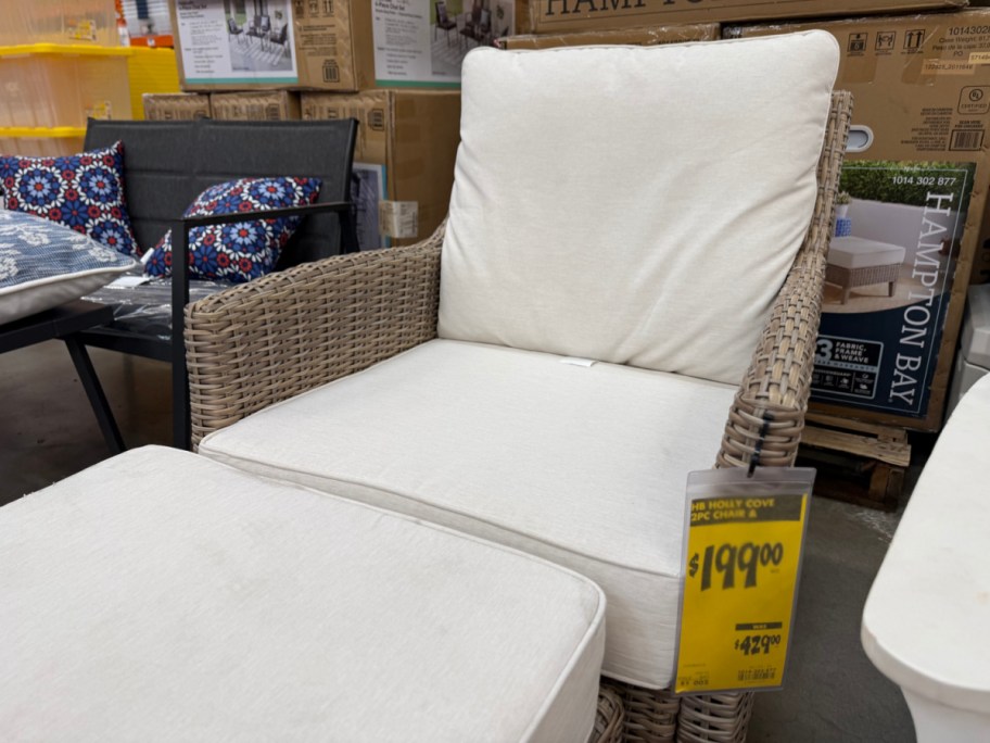 A wicker patio chair with cream cushions and a matching ottoman on display in a store with a yellow $199.00 clearance tag attached.