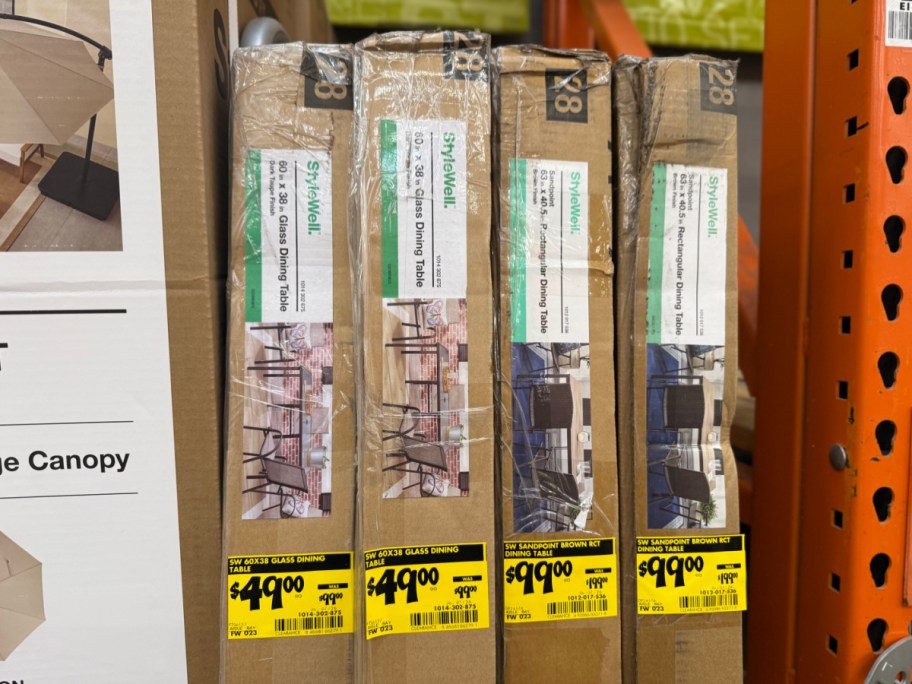 Four tall cardboard boxes for StyleWell patio dining tables standing upright in a store with yellow clearance price tags attached near the bottom.