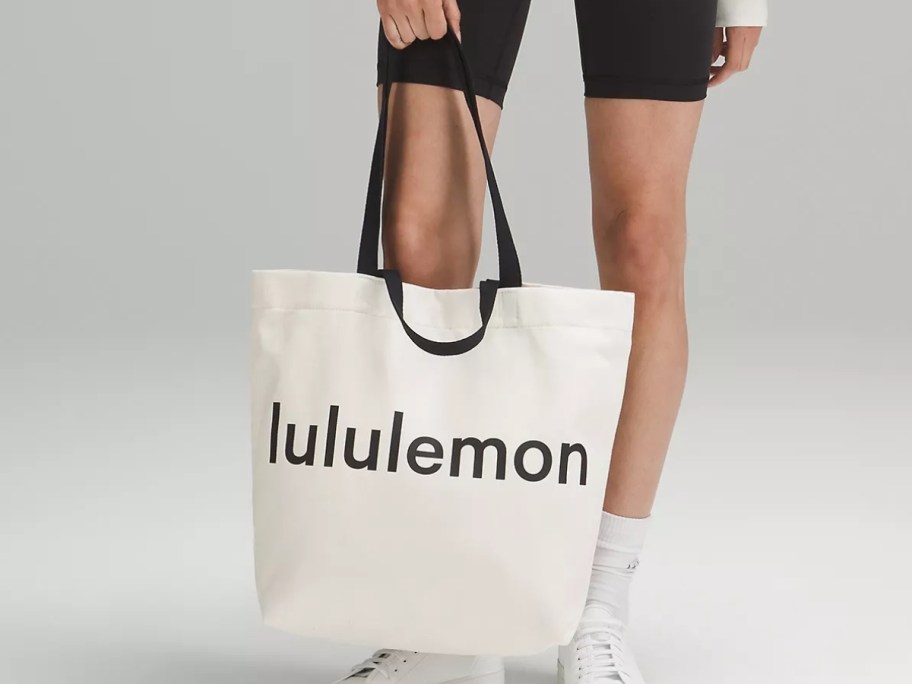 woman's legs and hand holding a canvas and black lululemon tote bag