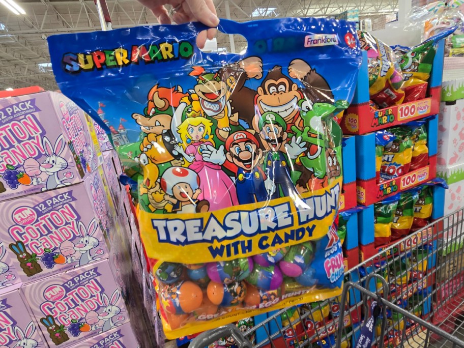 Bag of "Super Mario Treasure Hunt with Candy" held in hand, featuring colorful Mario characters. Candy aisle background with cotton candy boxes.