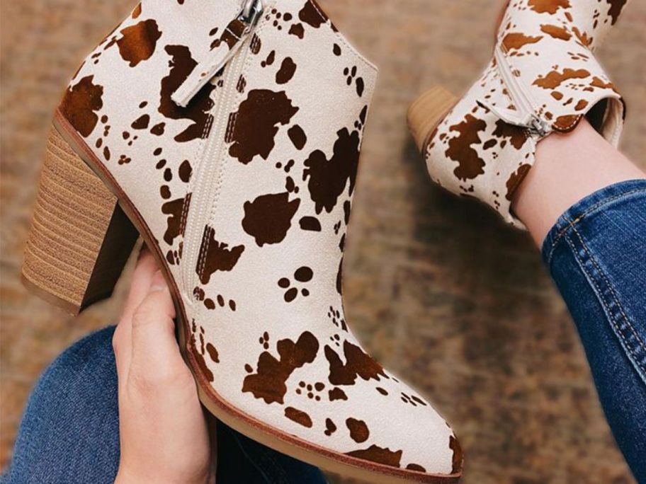 Hand holding a Maurices Cow Print Boot in left hand while wearing one on the right foot