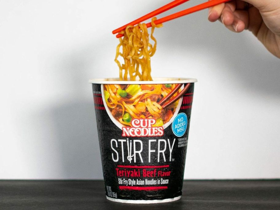 Nissin Stir Fry Noodles in Sauce Teriyaki Beef Flavor 3oz cup with noodles being taken out by hand with chop sticks on table