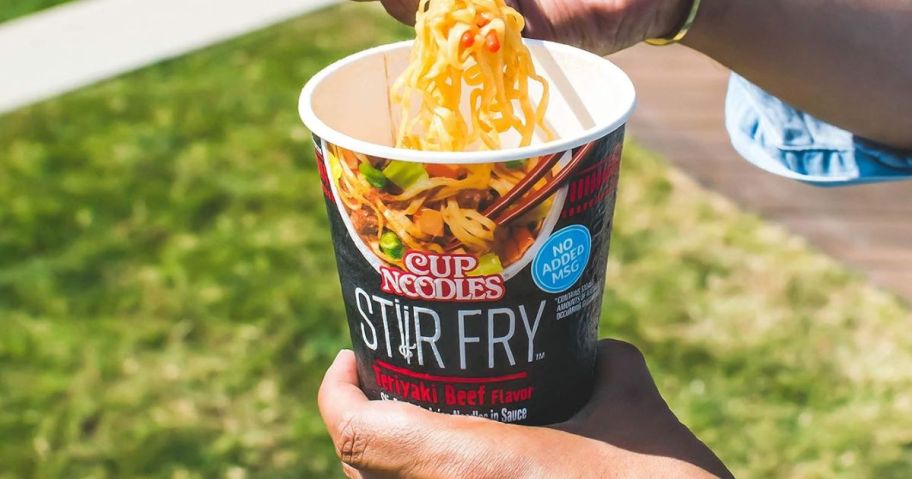 Nissin Cup Noodles Stir Fry Noodles in Sauce, Teriyaki Beef, 3 Ounce open with noodles being taken out of it by hand with chop sticks at a park