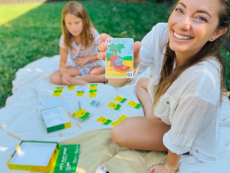 Sara smiling and holding a Play Nine card to the camera, with her daughter sitting behind her as they are playing the game on a blanket outdoors
