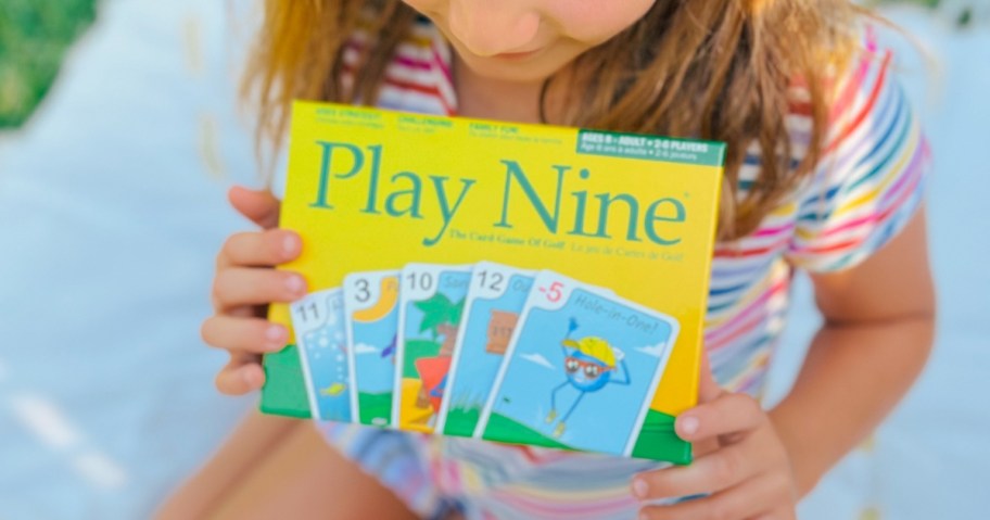 little girl holding a Play Nine Golf Card Game, she's sitting outside on a blanket