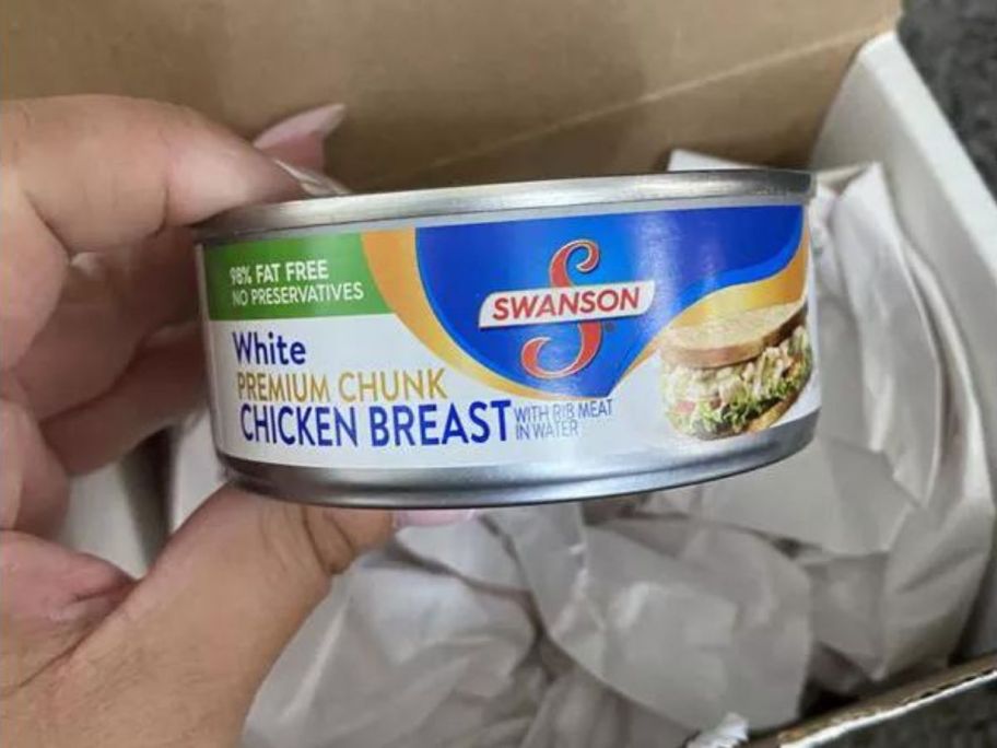 Swanson White Premium Chunk Canned Chicken Breast in Water, Fully Cooked Chicken, 12.5 OZ Can being taken out by hand out of a box