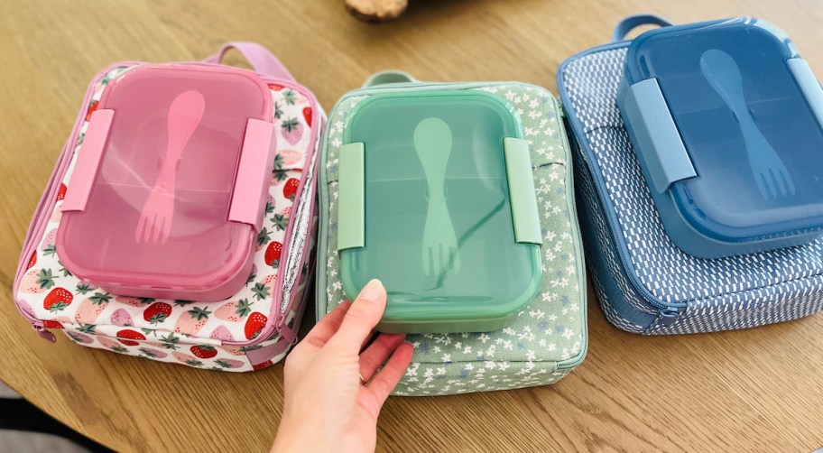 hand holding a green bento box on lunch box with other kids lunch box sets on wood table