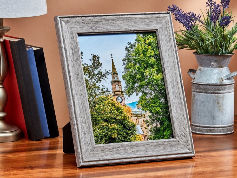 walmart picture frame showing a scenic image set on a table near books, a lamp, and a vase with flora