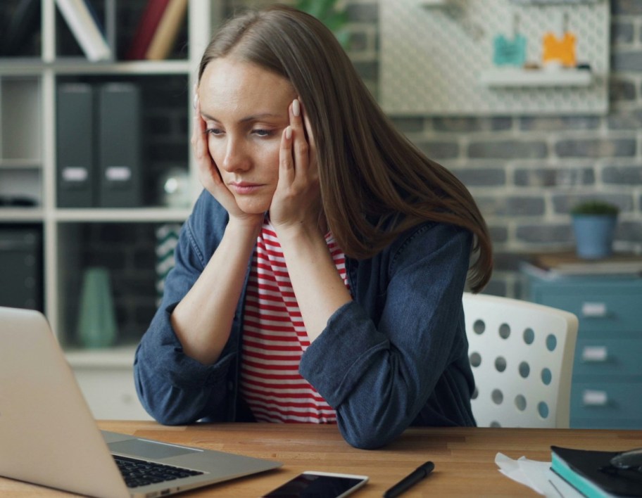 woman with hands on chin sitting at desk looking sad at laptop