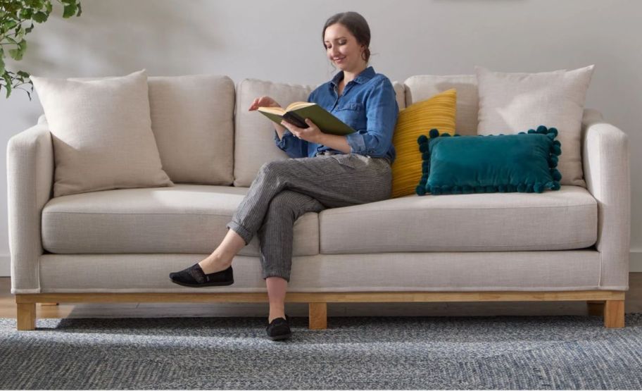 a woman sitting on an oatmeal colored sofa reading a book