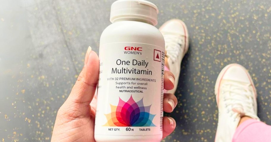 woman's hand holding a bottle of GNC Women's One Daily Multivitamin, her legs and feet below it