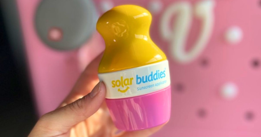 hand holding a pink and yellow Solar Buddies sunscreen applicator, pink Bogg bag in the Background