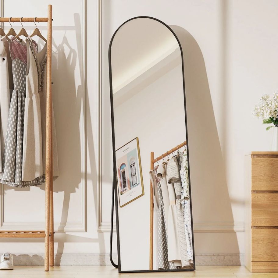 a black framed full length floor mirror next to a rack of clothes