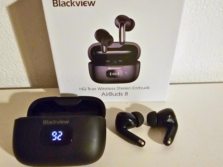 a pair of black wireless earbuds next to the charging case, a white box with the packaging they come in behind them