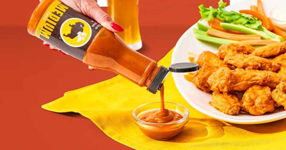 pouring a bottle of Buffalo Wild Wings Medium Buffalo Sauce with plate of wings behind it