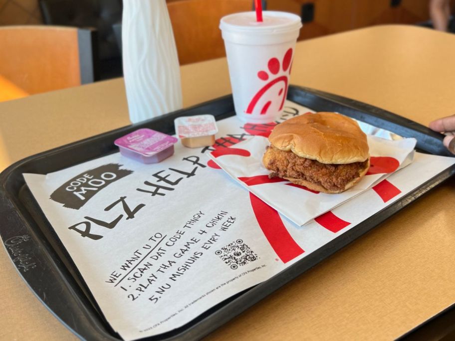 A tray of Chik Fil A food with a chicken sandwich and a drink on it with a paper liner Advertising their Code Moo Game