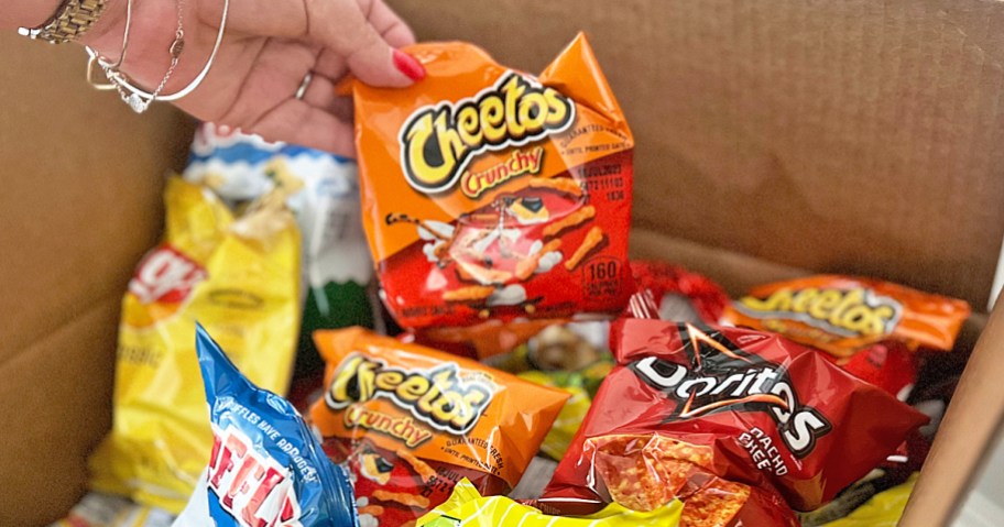 hand grabbing a bag of cheetos from a cardboard boxed filled with snacks
