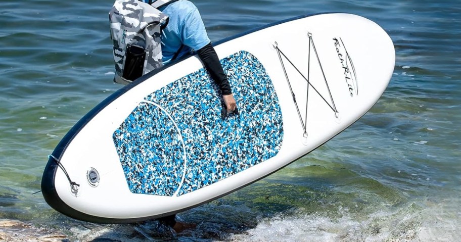 man carrying a large white and blue inflatable paddleboard into the ocean