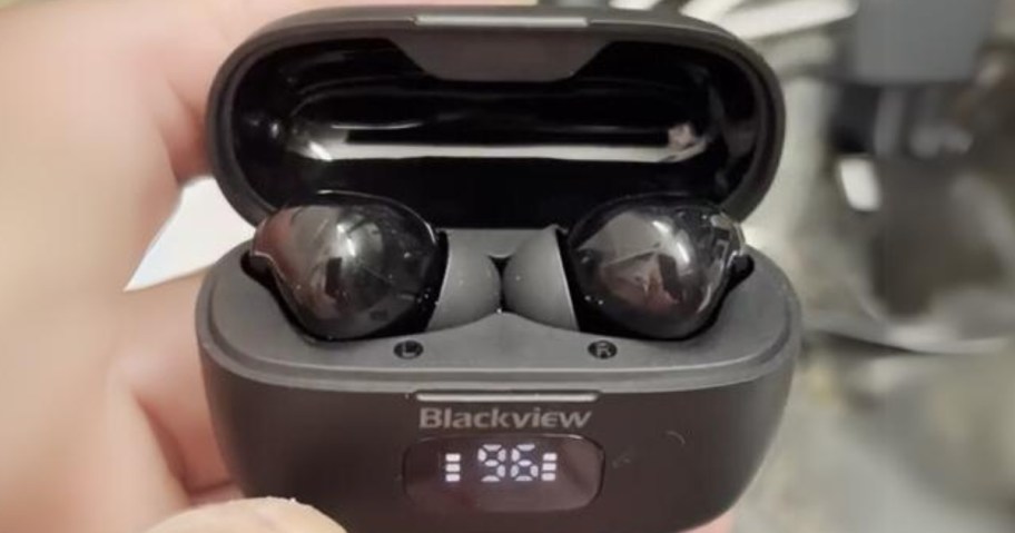 hand holding a a pair of black wireless earbuds in a black charging case that shows the battery level