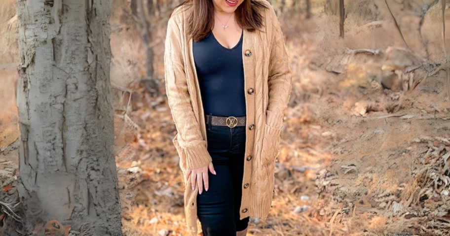 woman standing in the woods wearing a tan long cable knit cardigan with buttons with a navy top and jeans