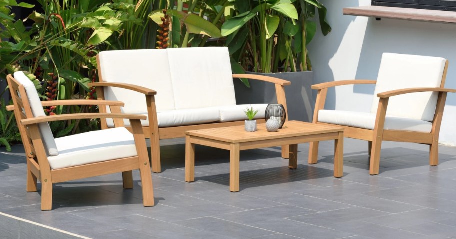 outdoor patio set in light wood with cream cushions, loveseat, 2 chairs and coffee table with plants behind them