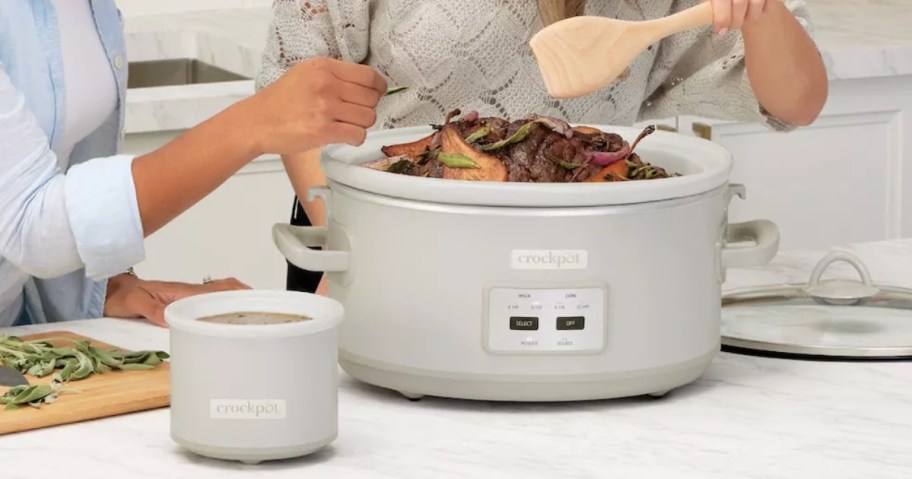 a mushroom cream color large Crockpot with a Little Dipper Crockpot on a kitchen island with person stirring the food in it and another person's hand adding seasoning